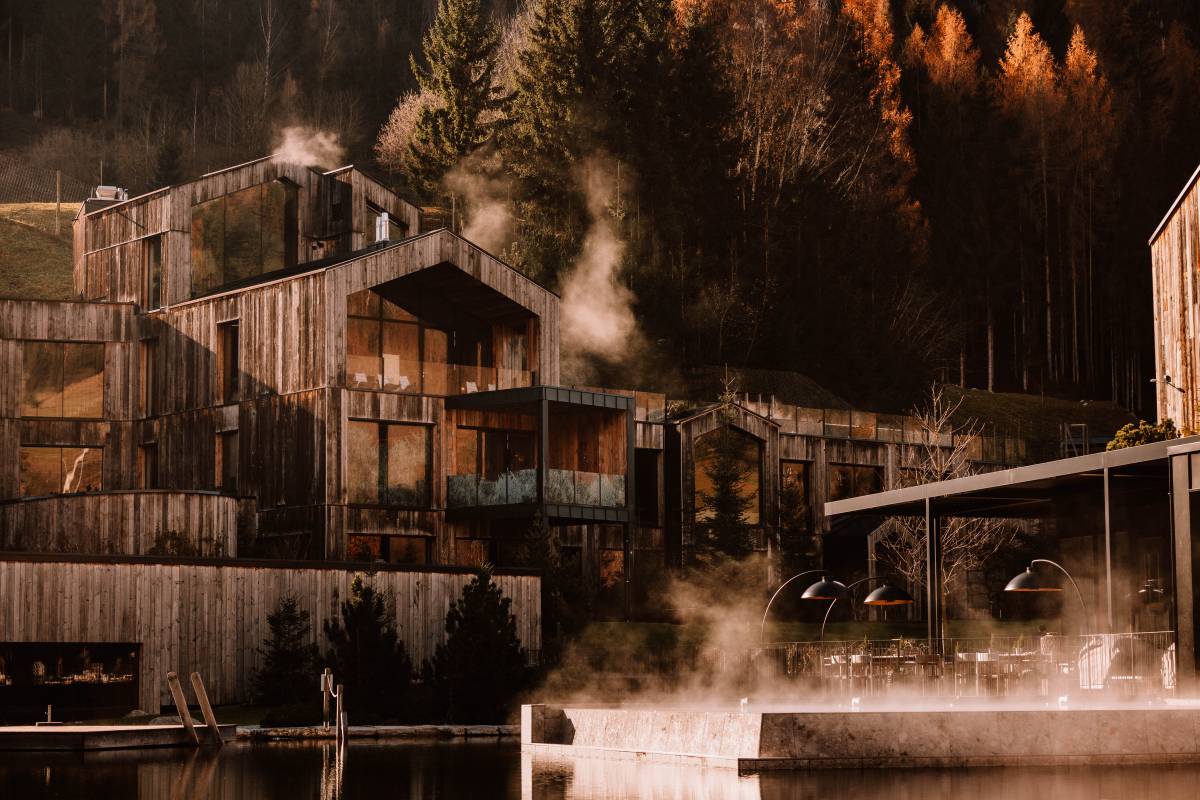 Naturhotel aus Holz im Herbst hinter einem See mit Wald im Hintergrund