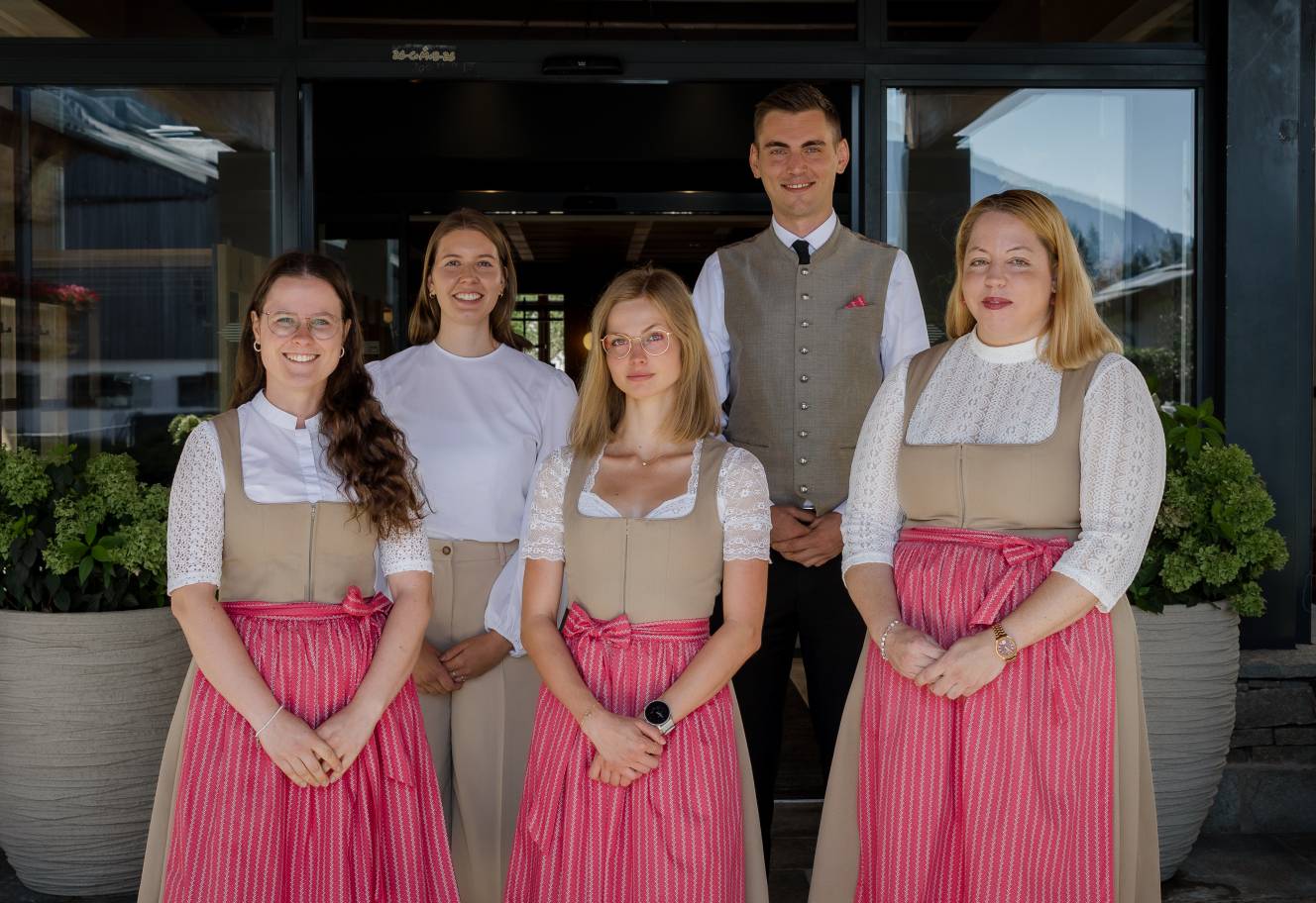 The five-member Glücksbringer team smiles in front of the hotel entrance. Three team members wear traditional dirndls, one wears a traditional vest, and one wears light-coloured work attire.