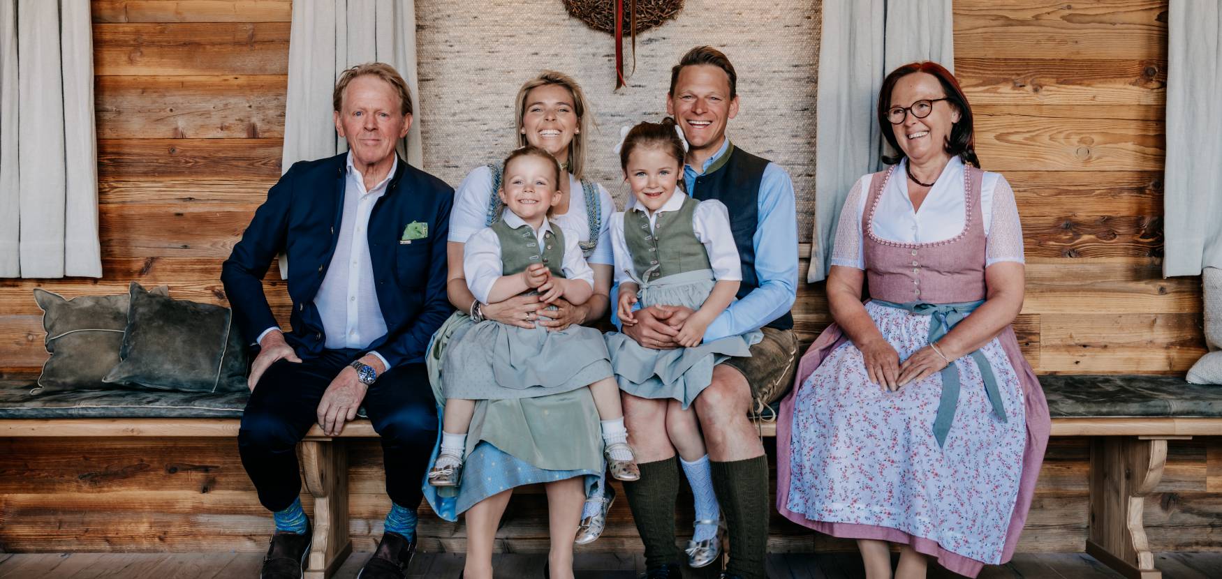 Drei Generationen einer Familie in traditioneller Tracht sitzen lachend auf einer Holzbank in einer gemütlichen Stube mit Holzwänden. Im Hintergrund hängt ein dekorativer Kranz an der Wand. Die familiäre Atmosphäre und die alpine Einrichtung spiegeln die herzliche Gastfreundschaft und die regionale Verbundenheit wider.