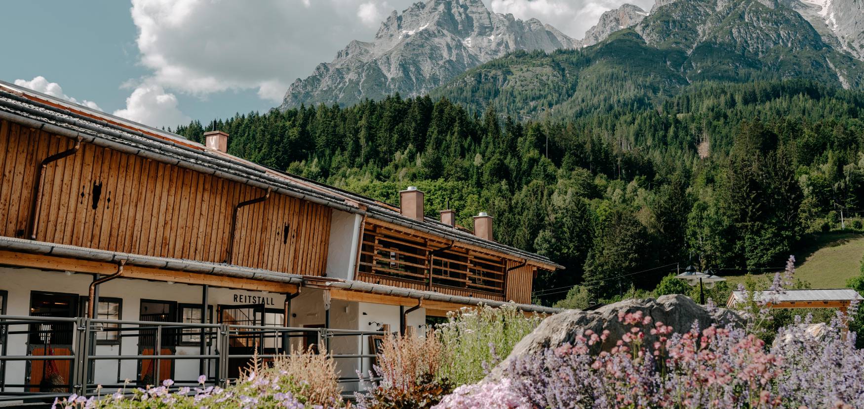 Reitstall mit Blumen und Bergpanorama im Sommer – Außenansicht.