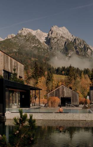 Moderne Wellnessanlage mit Infinity-Pool und Naturbadeteich, umgeben von herbstlichen Bäumen und Blick auf die imposanten, schneebedeckten Alpen. Eine Person genießt entspannte Momente im warmen Wasser, während im Hintergrund majestätische Berge und ein leichter Morgennebel Sehnsucht nach Erholung in der Natur wecken.