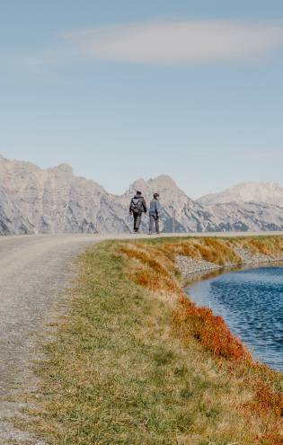Zwei Wanderer beim Wandern in Leogang entlang eines idyllischen Bergsees, umgeben von herbstlichen Almwiesen und beeindruckendem Alpenpanorama.