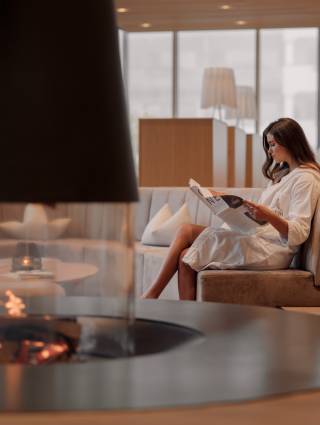 Woman in a bathrobe sitting in the lounge area of Naturhotel Forsthofgut, reading a newspaper in front of a modern fireplace.