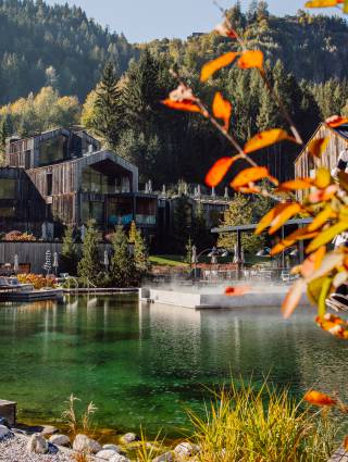 Naturbadesee mit dampfendem Außenpool, moderner Holzarchitektur und herbstlich gefärbten Wäldern in der Wellnesslandschaft des Forsthofguts.
