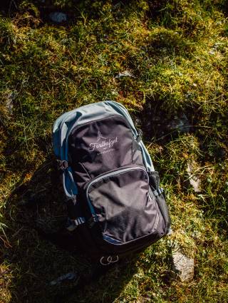 Schwarz-grauer Wanderrucksack mit Forsthofgut-Logo auf einer sonnigen Almwiese in Leogang, bereit für eine Wanderung in den Salzburger Alpen.