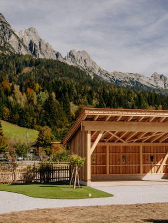 Der gepflegte Innenhof unseres Reitstalls mit grünem Rasen, Zierbäumen und Naturstein. Dahinter die Reithalle mit Holzfassade vor herbstlichem Wald und Bergkulisse.