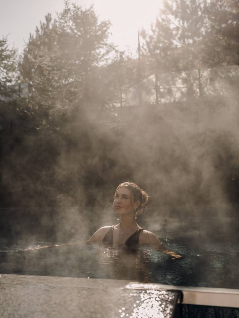 A woman relaxes in the steaming outdoor pool, surrounded by soft morning light and pine trees. Rising steam and the natural setting evoke tranquility, relaxation, and pure wellness in the Austrian Alps.