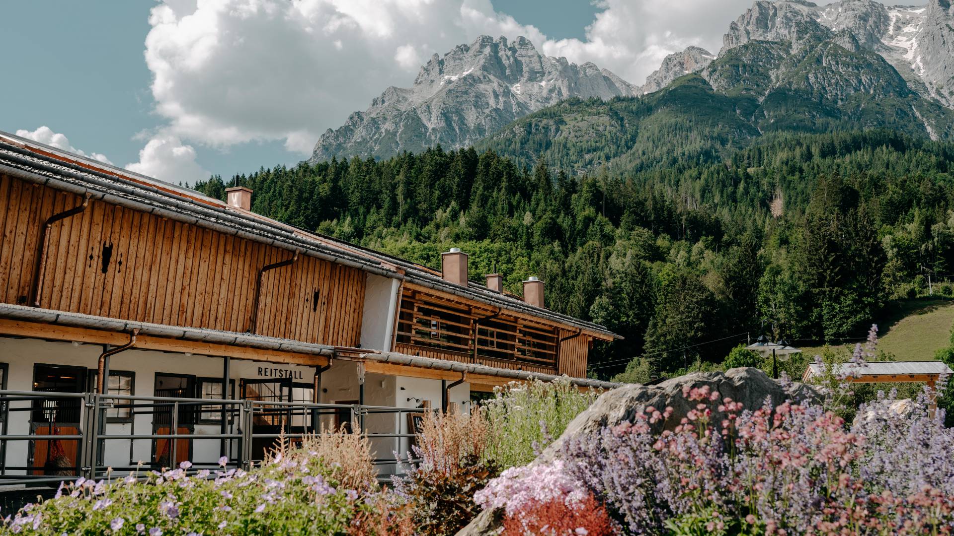 Reitstall mit Blumen und Bergpanorama im Sommer – Außenansicht.