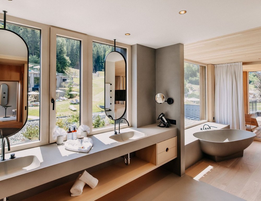 Bathroom with a freestanding bathtub, modern fixtures, and a suspended mirror. Behind it is a window wall overlooking the garden.
