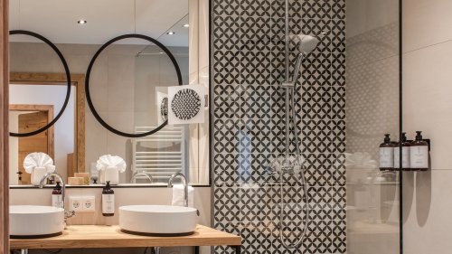 Badezimmer mit großem Spiegel, darüber zwei runde Spiegel. Die freistehenden Waschbecken stehen auf einer Armatur, direkt daneben befindet sich eine durchsichtige Duschwand.