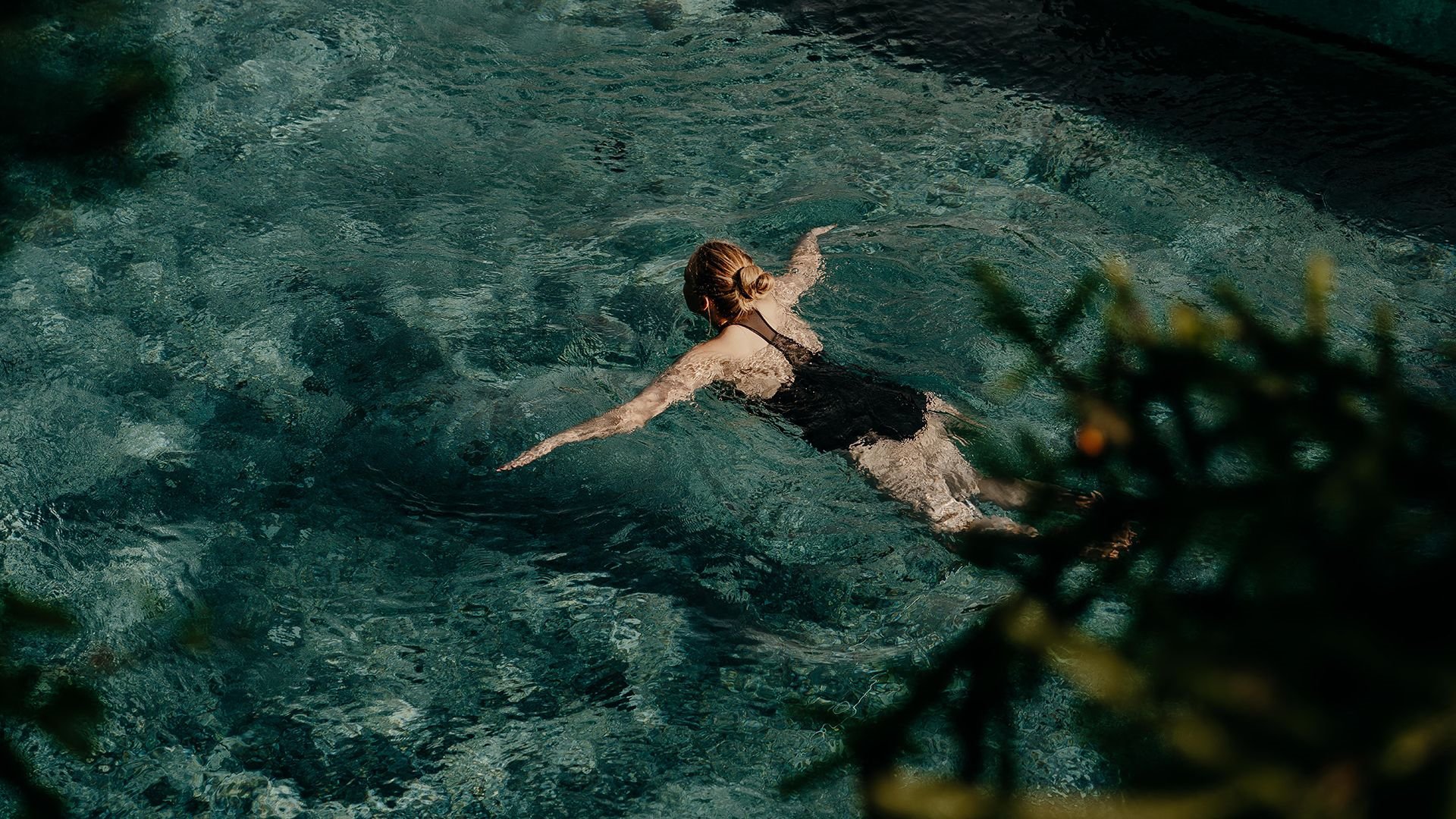 Woman swimming in the pool on a sunny day, with dappled light and shadow from nearby trees.