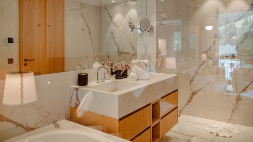 Elegantes Badezimmer der Familiensuite Bergbalance im Naturhotel Forsthofgut. Das Bild zeigt einen großzügigen Raum mit hellen Marmorwänden, modernem Doppelwaschtisch und stilvollen Holzelementen. Durch die Glasfront fällt Tageslicht in den Raum und eröffnet zugleich den Blick auf die Bergwelt. Warme Akzente, edle Materialien und dezente Beleuchtung schaffen eine exklusive Wellness-Atmosphäre im privaten Rückzugsort.