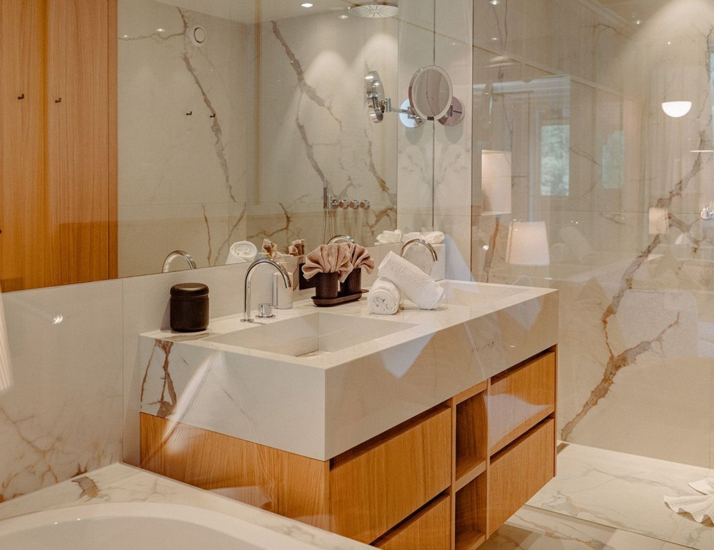 Elegant bathroom of the Family Suite Bergbalance at Naturhotel Forsthofgut. The image shows a spacious room with bright marble walls, a modern double washbasin, and stylish wooden details. Natural light enters through the glass front, offering a view of the surrounding mountains. Warm accents, refined materials, and subtle lighting create an exclusive spa-like atmosphere in this private retreat.