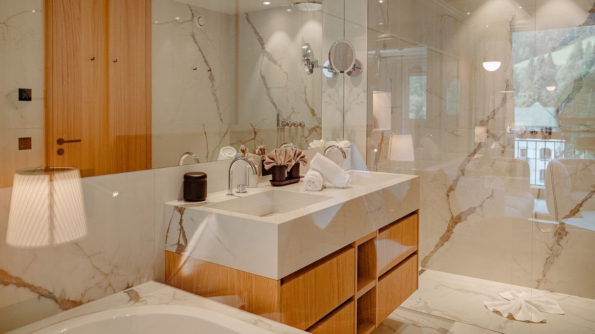 Elegant bathroom of the Family Suite Bergbalance at Naturhotel Forsthofgut. The image shows a spacious room with bright marble walls, a modern double washbasin, and stylish wooden details. Natural light enters through the glass front, offering a view of the surrounding mountains. Warm accents, refined materials, and subtle lighting create an exclusive spa-like atmosphere in this private retreat.