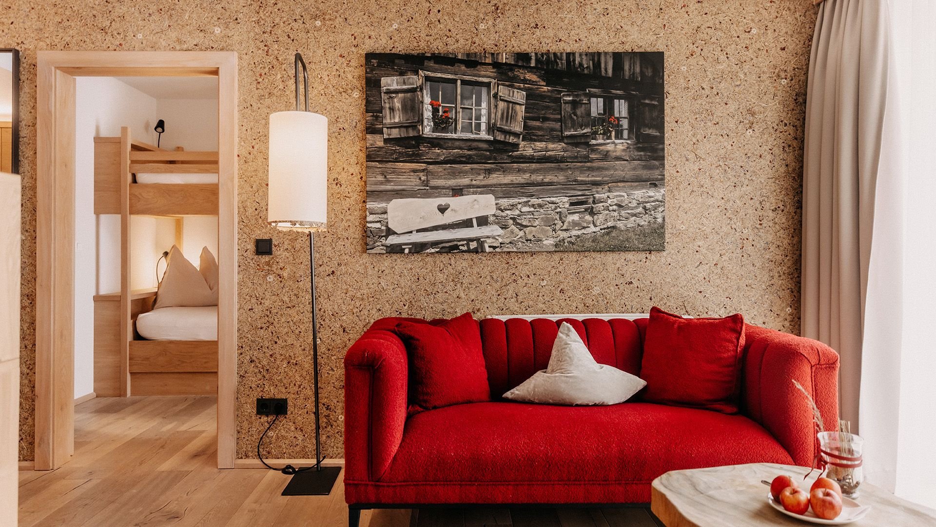 Section of a stylish furnished suite with a red couch, decorative pillows, and a black floor lamp. In the background, a children's room with a bunk bed is visible through an open door.