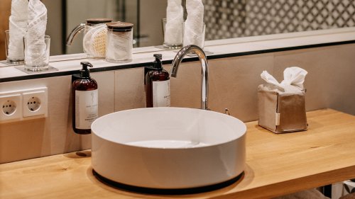 Ausschnitt aus einem stilvollen Hotelbadezimmer mit einem runden Aufsatzwaschbecken auf einer eleganten Armatur; daneben stehen hochwertige Hotelkosmetikprodukte.