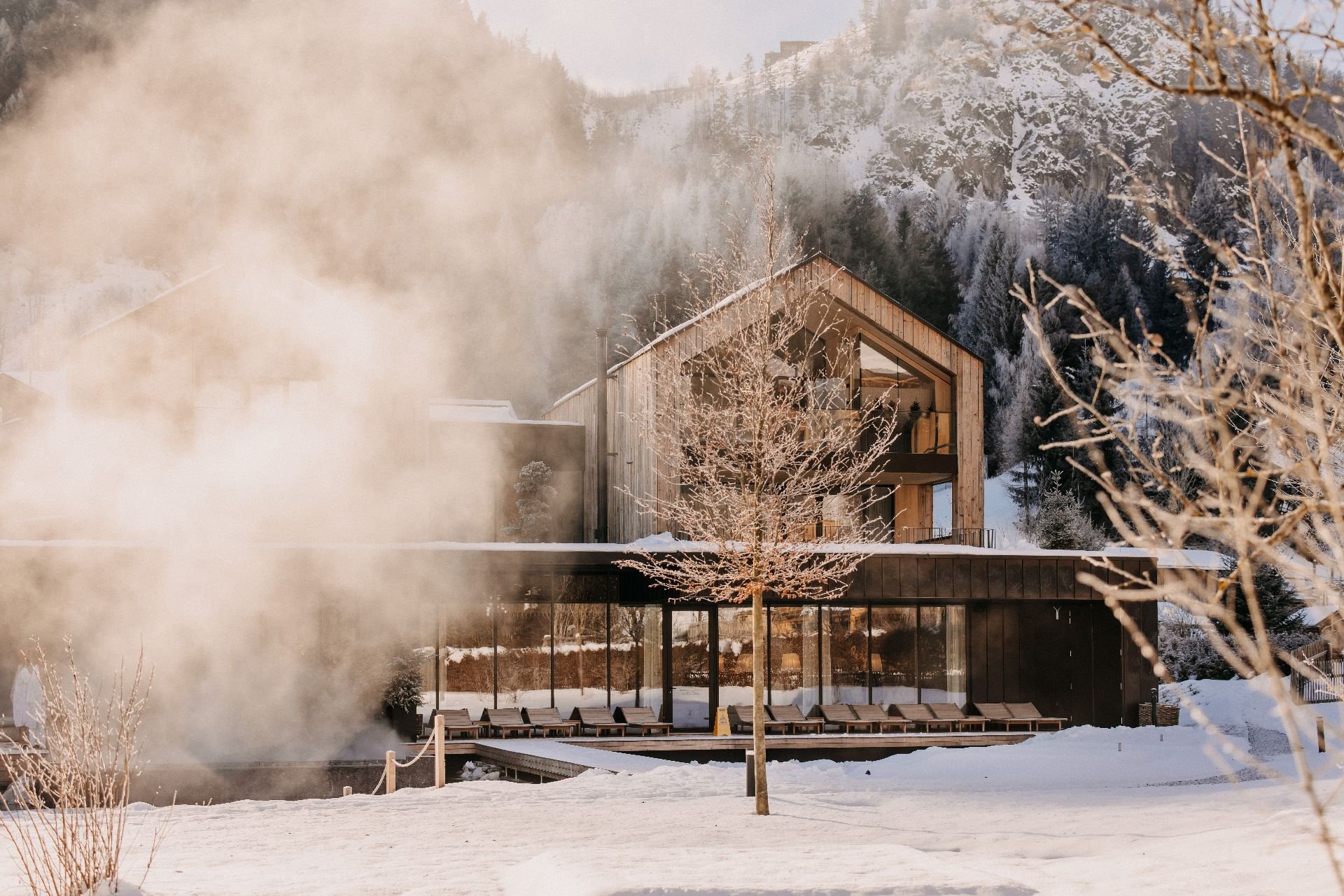 The lakeside house at Forsthofgut in winter, featuring a snow-covered landscape, a steaming natural swimming lake and contemporary wooden architecture set against an alpine mountain backdrop in Leogang.