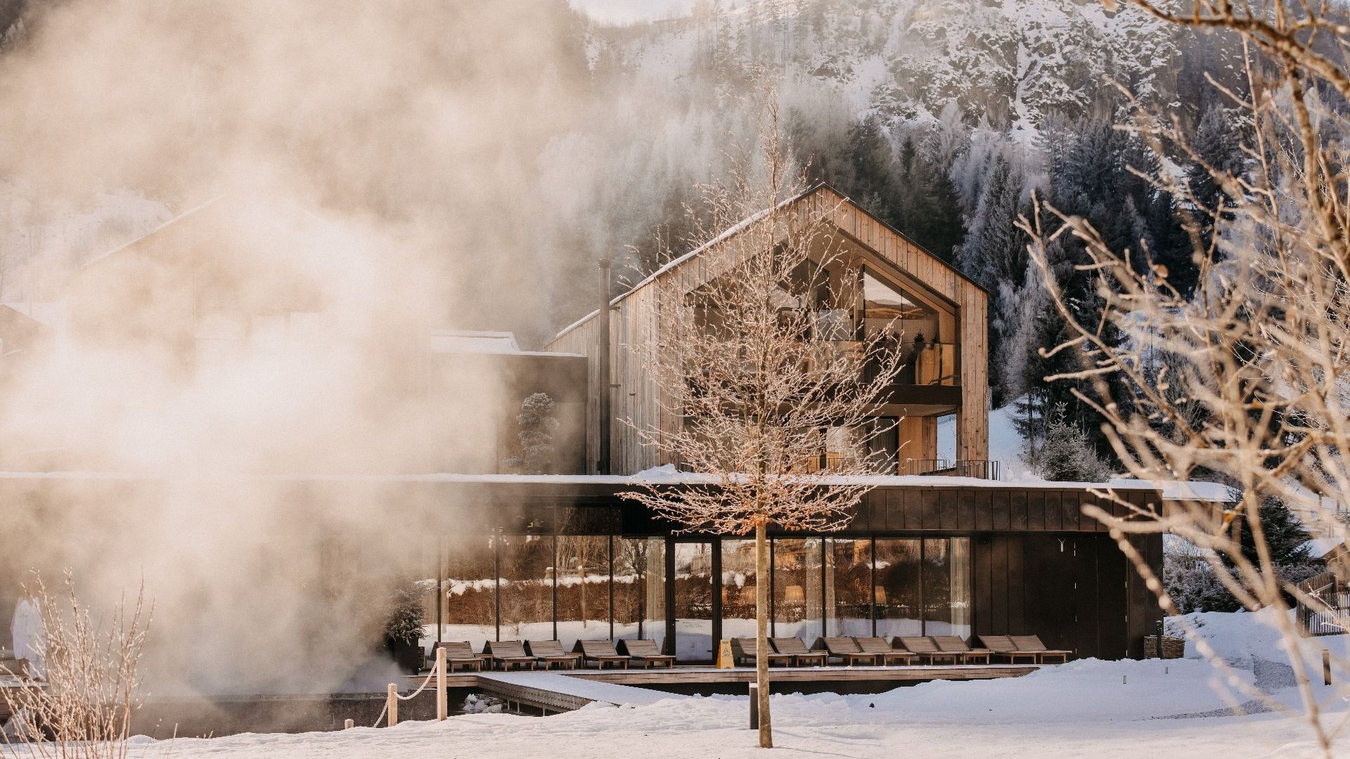 The lakeside house at Forsthofgut in winter, featuring a snow-covered landscape, a steaming natural swimming lake and contemporary wooden architecture set against an alpine mountain backdrop in Leogang.