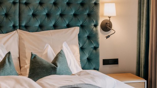 Ausschnitt eines gemütlichen Bettes mit weißer Bettwäsche, türkise gepolsterter Rückenlehne und türkise Zierkissen. Daneben steht ein Nachttisch mit einer an der Wand montierten Nachtlampe.