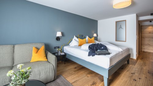 Helles Naturzimmer Landidyll mit Doppelbett, gemütlichem Sofa und Holzboden vor blauer Wand.