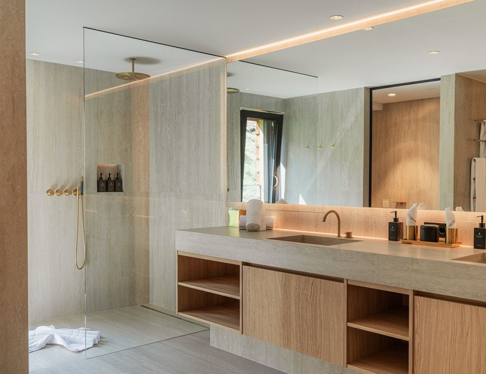 Modern bathroom in natural sand and wooden tones with a spacious walk-in rain shower, elegant gold fixtures, and a long vanity with two sinks. Subtle decorations such as soap dispensers and towels enhance the luxurious atmosphere, with a view of greenery through the window.