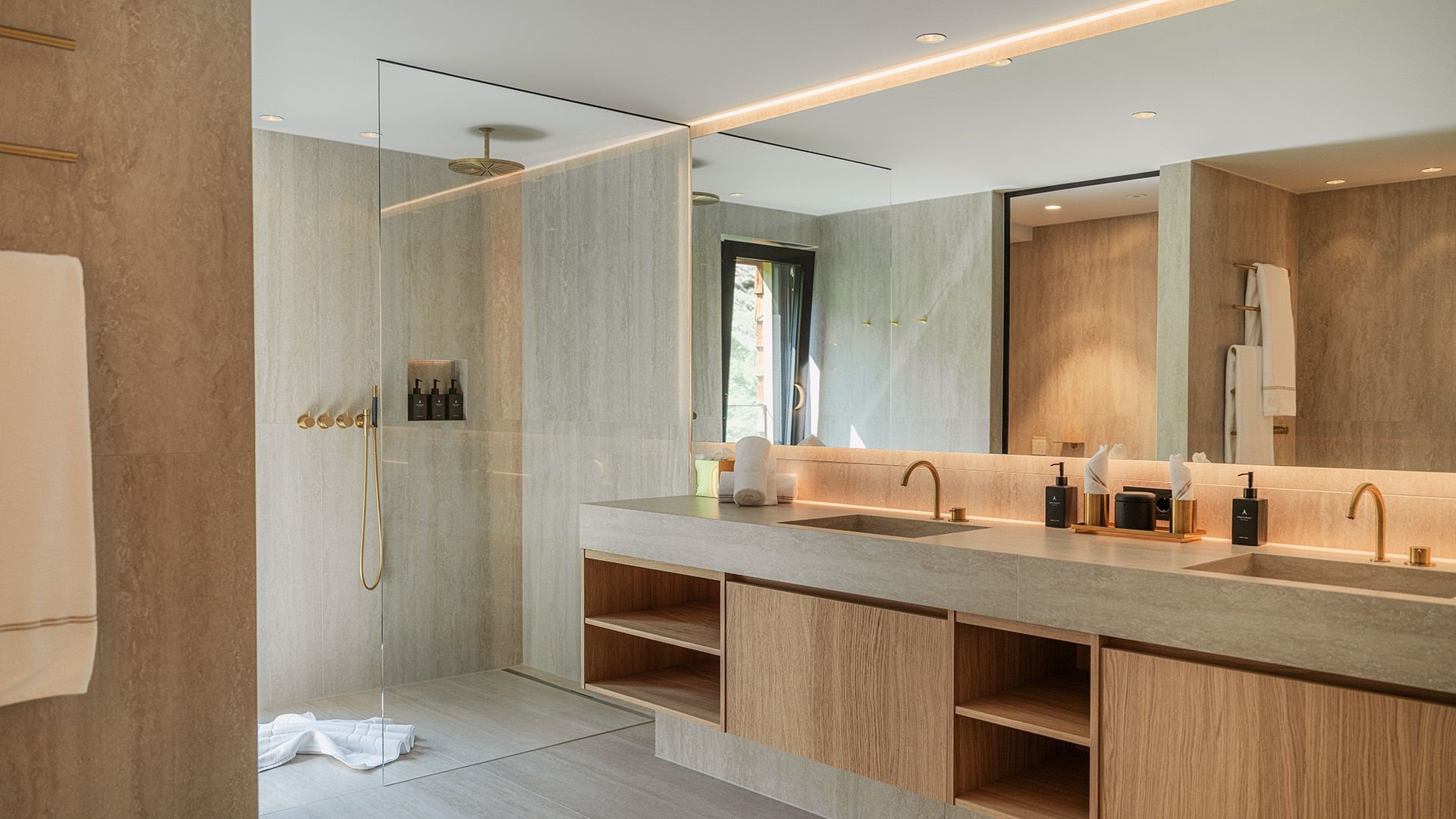 Modern bathroom in natural sand and wooden tones with a spacious walk-in rain shower, elegant gold fixtures, and a long vanity with two sinks. Subtle decorations such as soap dispensers and towels enhance the luxurious atmosphere, with a view of greenery through the window.