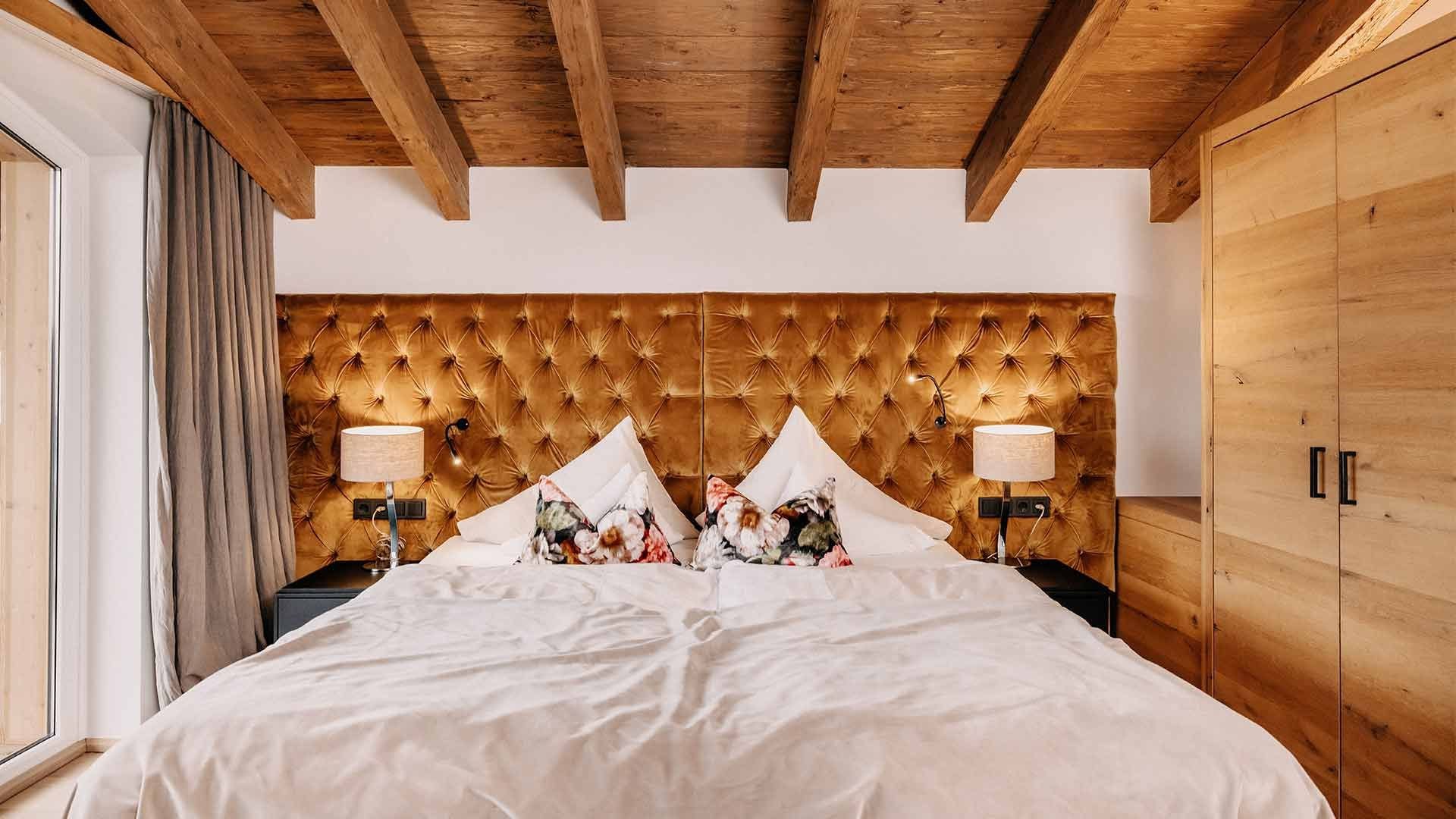 Großes Bett mit goldfarbener Kopfteilpolsterung und Zierpolstern. Rechts und links stehen jeweils Nachttische mit Nachttischlampen. Auf der rechten Seite befindet sich ein großer Schrank und auf der linken Seite ein Fenster mit grauen Vorhängen.