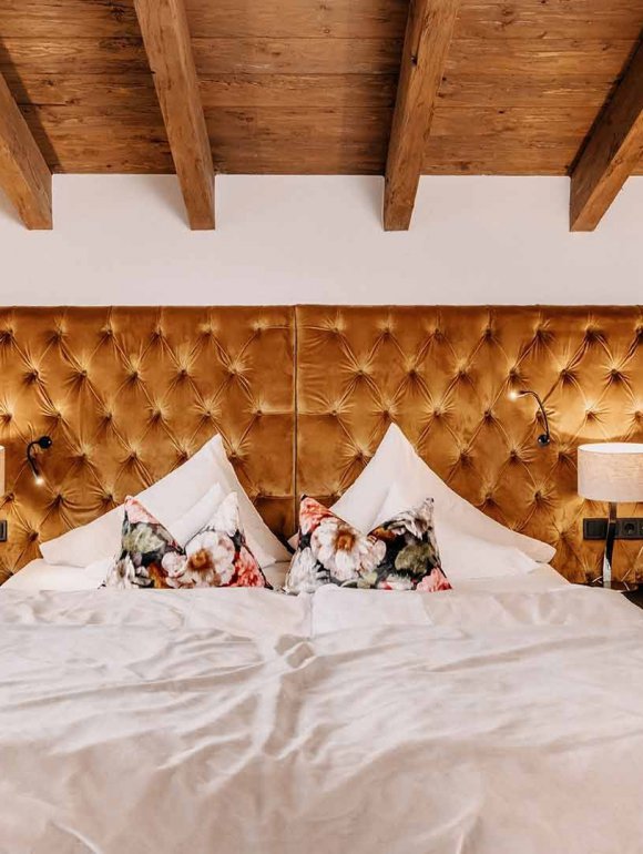 A large bed with a gold-colored upholstered headboard and decorative pillows. On both sides, there are nightstands topped with bedside lamps. On the right side, a large wardrobe is visible, while on the left side, there is a window with gray curtains.