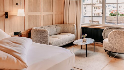 Modern eingerichtetes Zimmer. In der linken Ecke sieht man einen Teil eines gemütlichen Bettes. Außerdem gibt es eine gemütliche Sitzecke mit Couch und Sessel. In der Mitte steht ein Tisch. Die Wand ist mit hellem Holzinterior verziert.