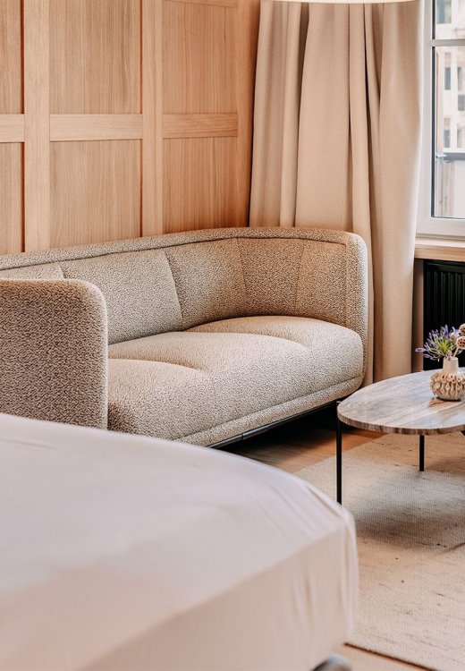 Modernly furnished room. In the left corner, part of a cozy bed is visible. There is also a comfortable seating area with a couch and an armchair. A table is positioned in the middle. The wall is decorated with elegant wood paneling.