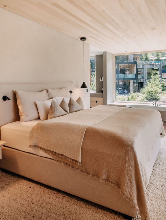 Room with a large, cozy bed, a nightstand with hanging bedside lamps beside it. Straight ahead is a large window overlooking the garden.