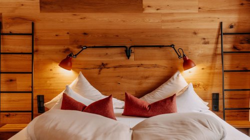 Gemütliches Doppelbett mit roter Akzentbeleuchtung und roten Zierkissen, vor einer rustikalen Holzvertäfelung mit integrierten schwarzen Metall-Elementen im modernen Alpenstil.