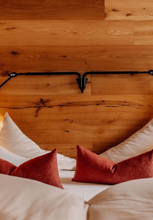 Inviting double bed with red accent pillows and matching red reading lamps, set against a rustic wooden wall with black metal frame elements in a modern Alpine design.