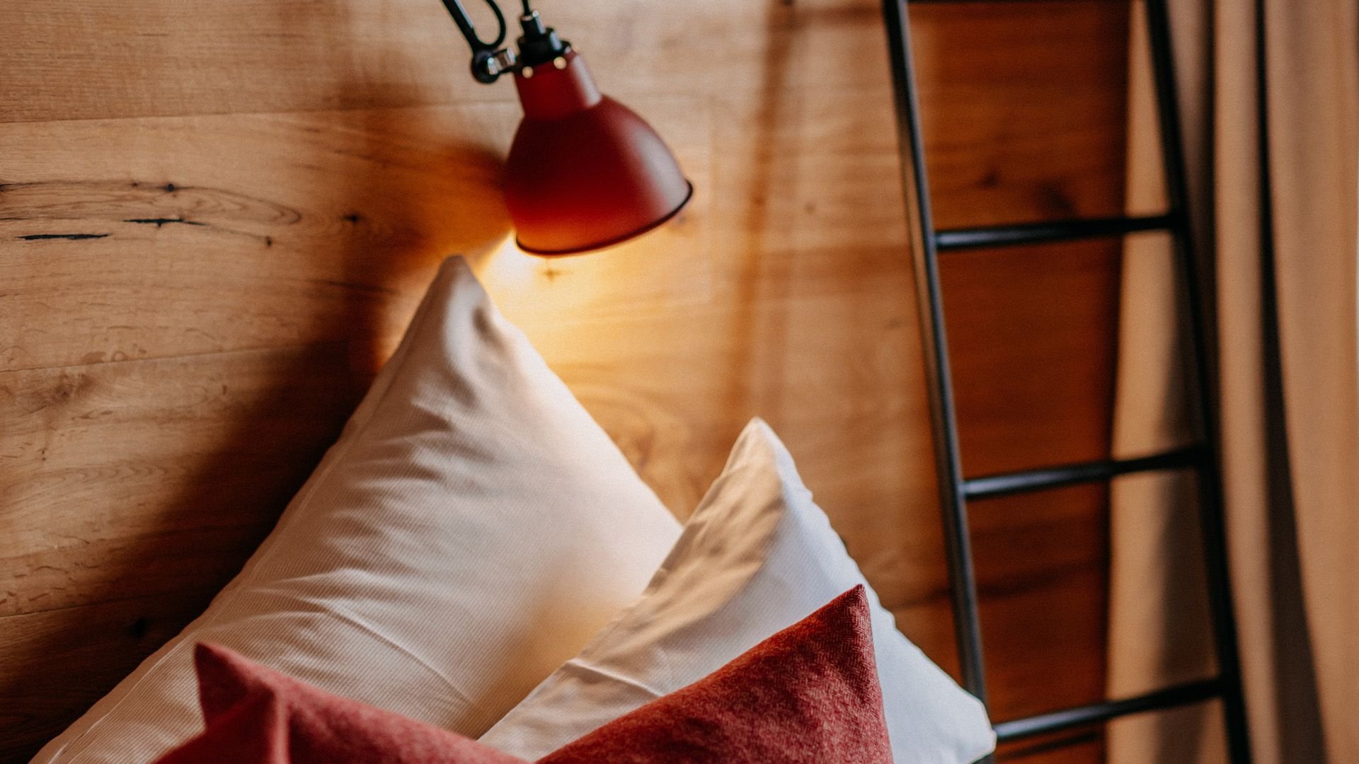 Close-up of a cozy hotel bed with white pillows, red decorative cushions, and a red wall-mounted lamp against a natural wooden wall.