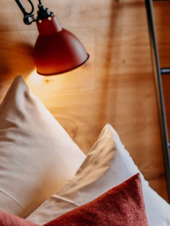 Close-up of a cozy hotel bed with white pillows, red decorative cushions, and a red wall-mounted lamp against a natural wooden wall.