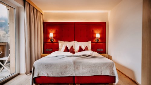 Cozy bed with white bedding, red accents, and a red upholstered headboard. On both sides, there is a nightstand with wall-mounted bedside lights above.