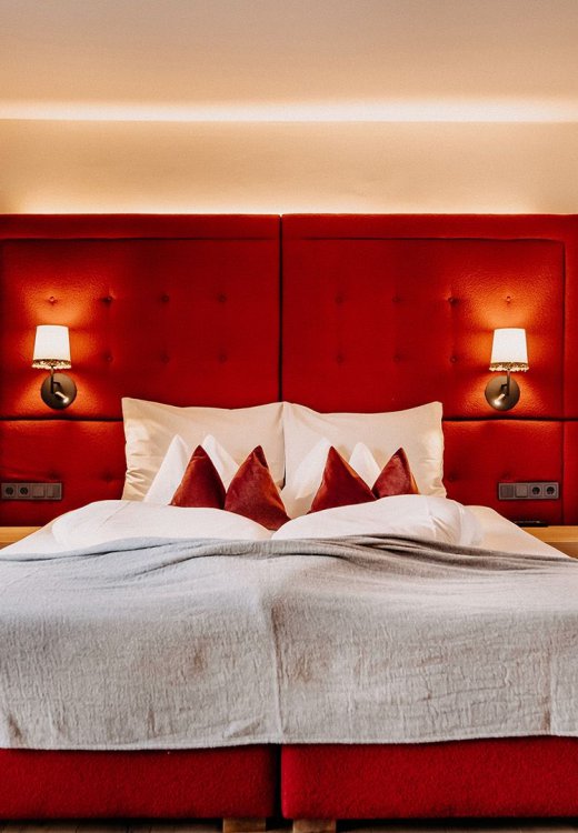 Cozy bed with white bedding, red accents, and a red upholstered headboard. On both sides, there is a nightstand with wall-mounted bedside lights above.