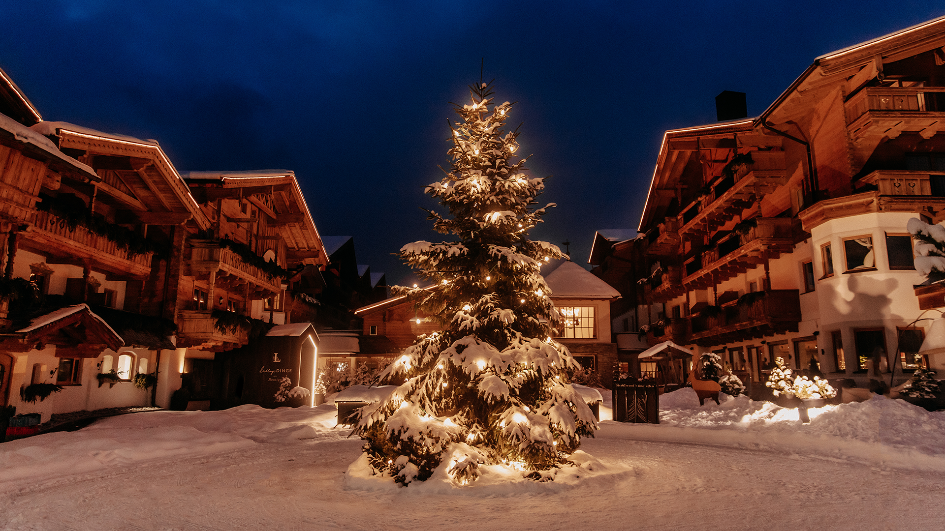 A Christmas tree surrounded by atmospheric lights in the snowy courtyard of the Naturhotel Forsthofgut at dusk.