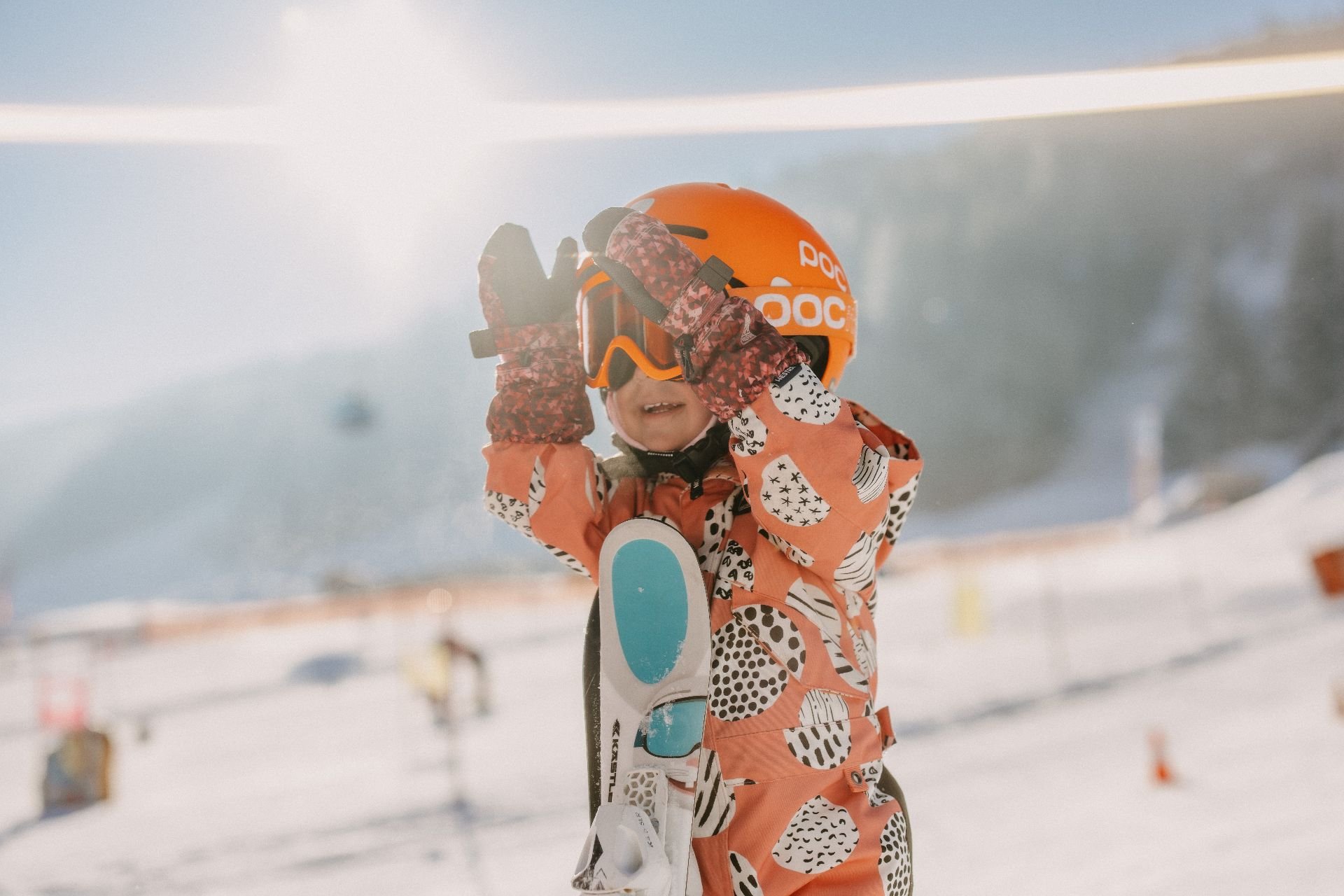 A child holding skis close to the body and playfully clapping hands in front of the face, with a sunny ski children’s area and alpine winter scenery in Leogang.