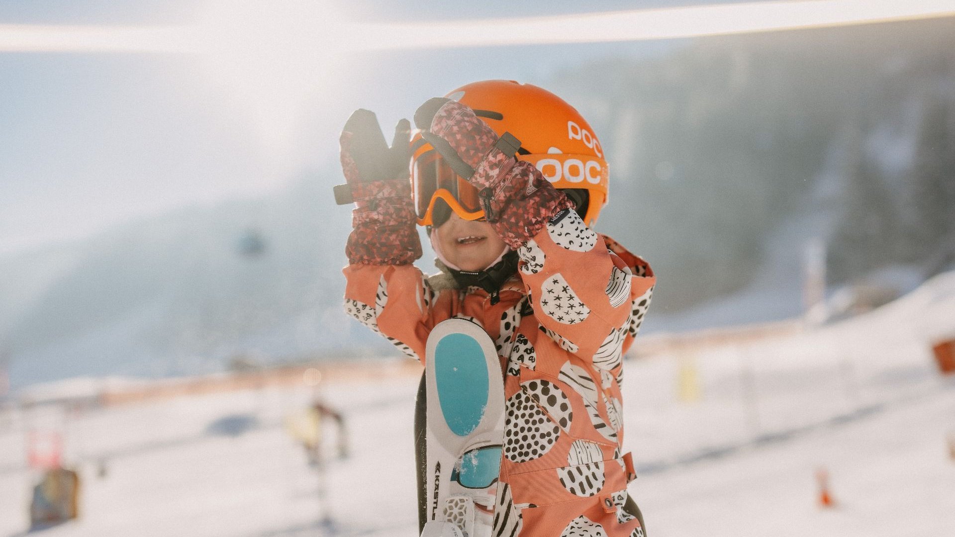 A child holding skis close to the body and playfully clapping hands in front of the face, with a sunny ski children’s area and alpine winter scenery in Leogang.