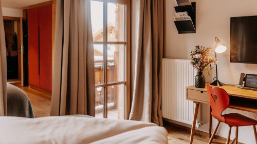 Teilansicht eines Hotelzimmers mit sichtbarer Bettkante im Vordergrund und einem Schreibtisch mit einem Stuhl im Hintergrund.