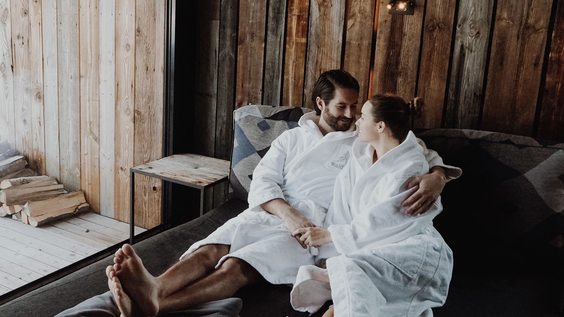 Couple in white bathrobes sits closely together on a lounge chair, smiling at each other.