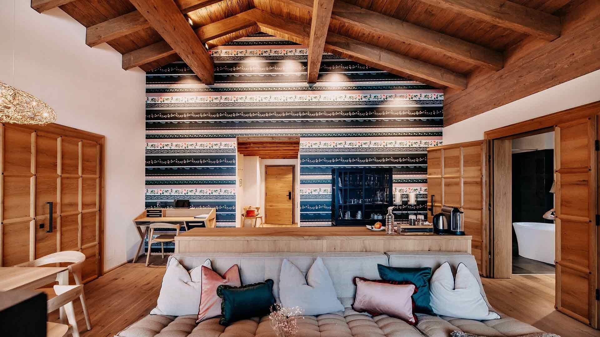 Spacious living area with a cozy couch and many wooden elements such as floors, cabinets, and doors. Behind it, there is a table with chairs and a sideboard with a coffee machine. On the right side, a freestanding bathtub is visible through the doorway.