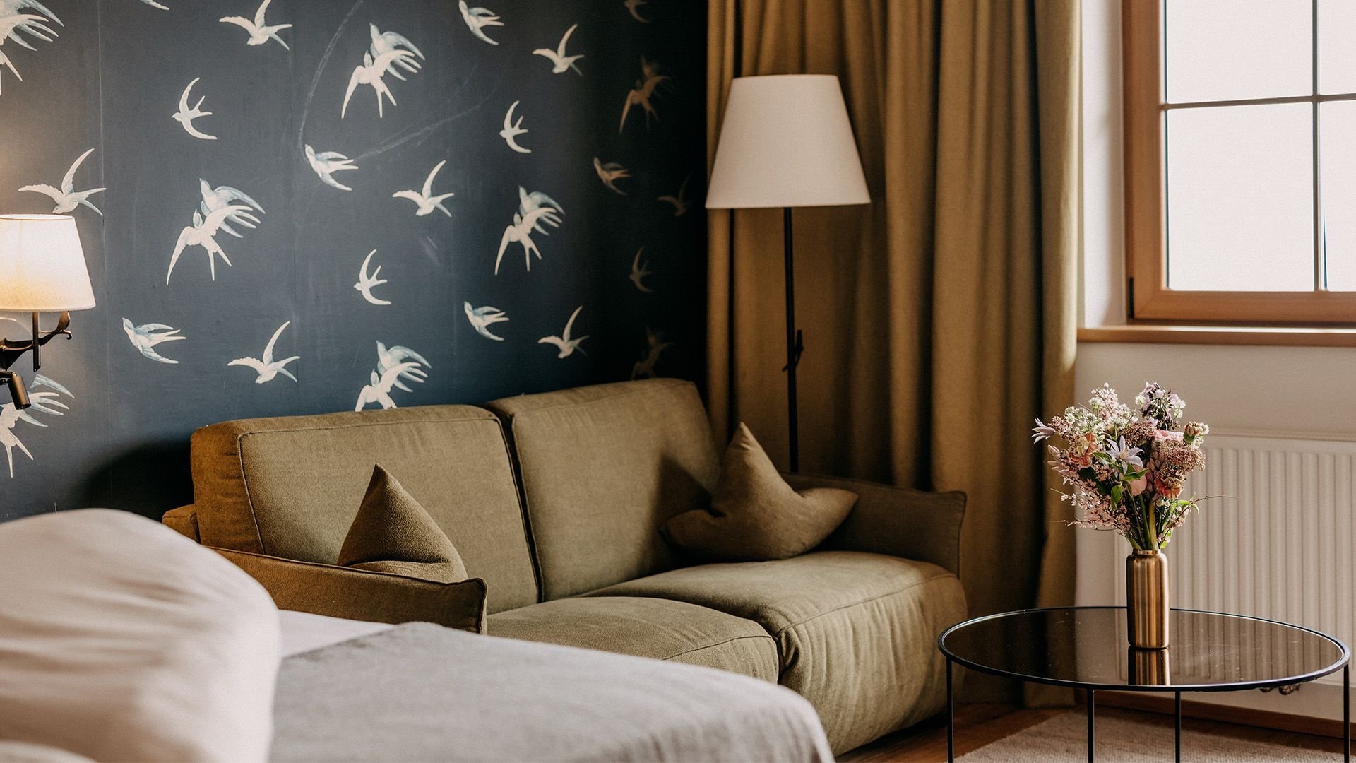 In the left corner, part of a cozy bed is visible. Behind it, there is an olive green couch in front of a wall with swallow-patterned wallpaper.