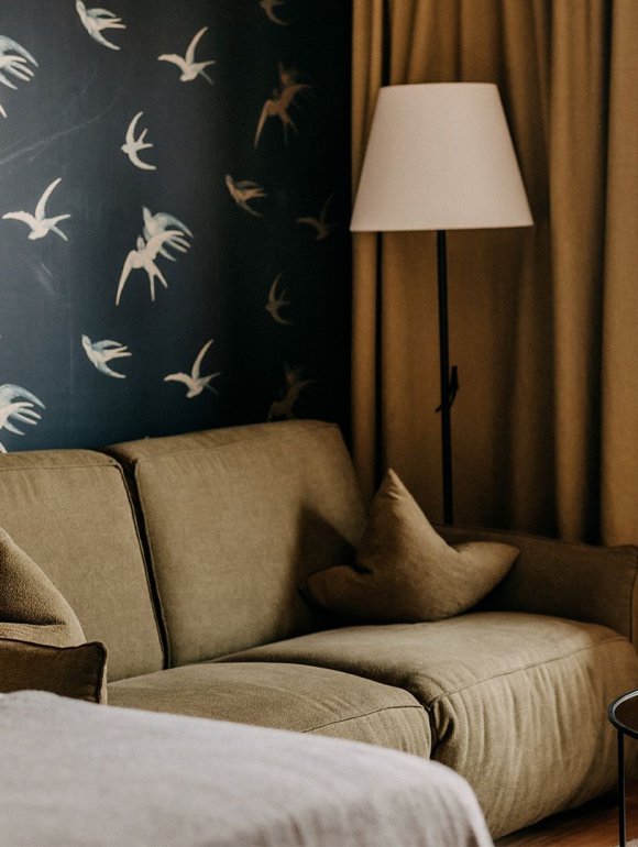 In the left corner, part of a cozy bed is visible. Behind it, there is an olive green couch in front of a wall with swallow-patterned wallpaper.
