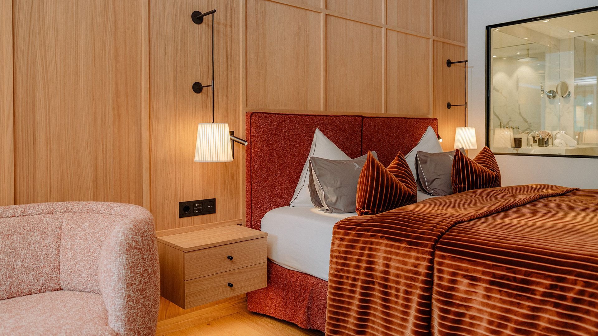 The stylishly furnished Family Suite Bergbalance at Naturhotel Forsthofgut. The image shows a cozy bedroom with elegant wood design, a large double bed with a deep red velvet headboard, refined decorative pillows, and soft lighting. A large glass wall provides a view into the modern bathroom, while warm colors, soft velvet accents, and premium materials create a welcoming and luxurious atmosphere.