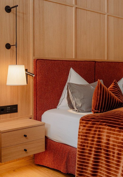 The stylishly furnished Family Suite Bergbalance at Naturhotel Forsthofgut. The image shows a cozy bedroom with elegant wood design, a large double bed with a deep red velvet headboard, refined decorative pillows, and soft lighting. A large glass wall provides a view into the modern bathroom, while warm colors, soft velvet accents, and premium materials create a welcoming and luxurious atmosphere.