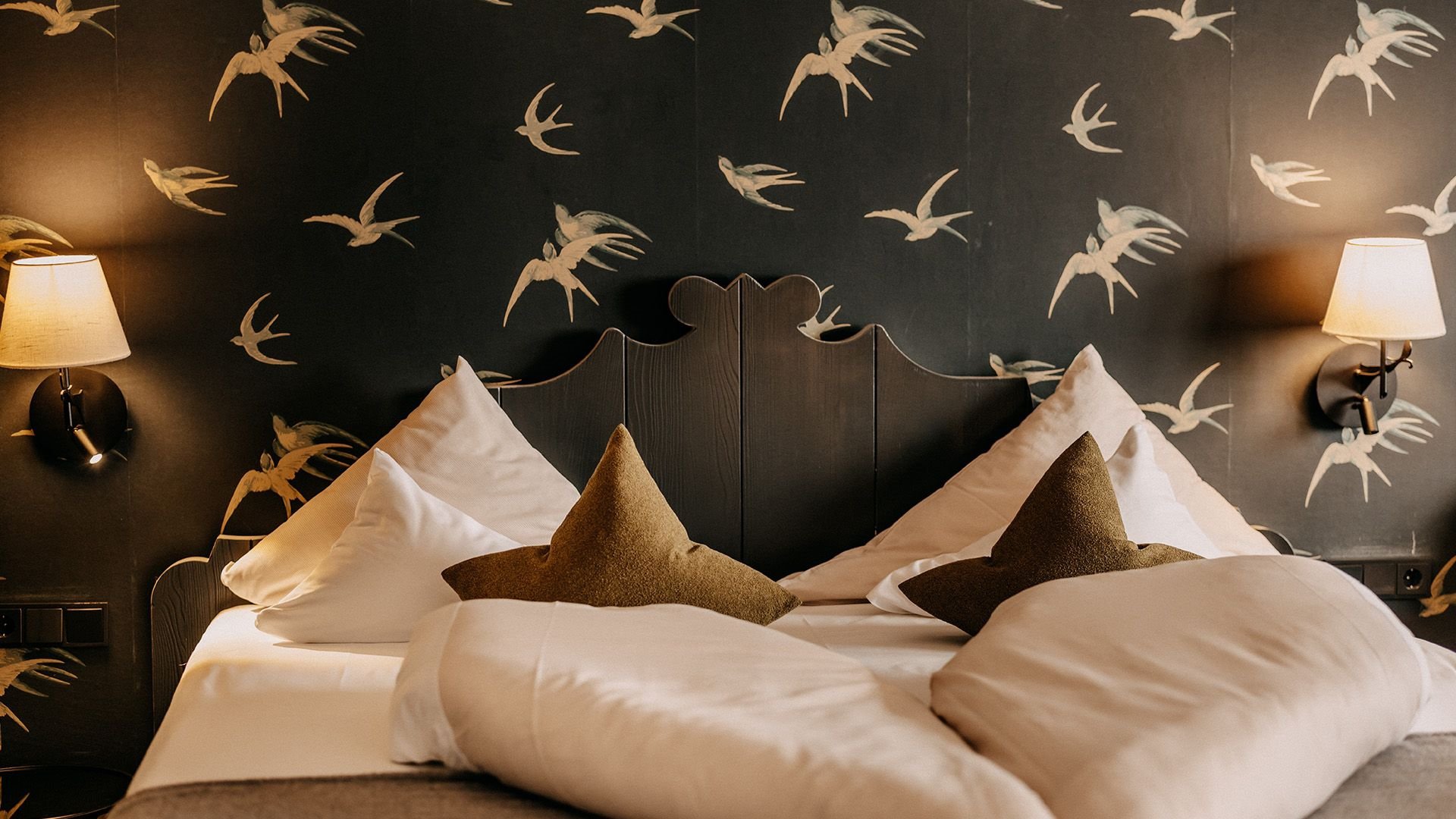 Close-up of a cozy bed with white bedding and an olive green upholstered headboard, with a swallow-patterned wallpaper.