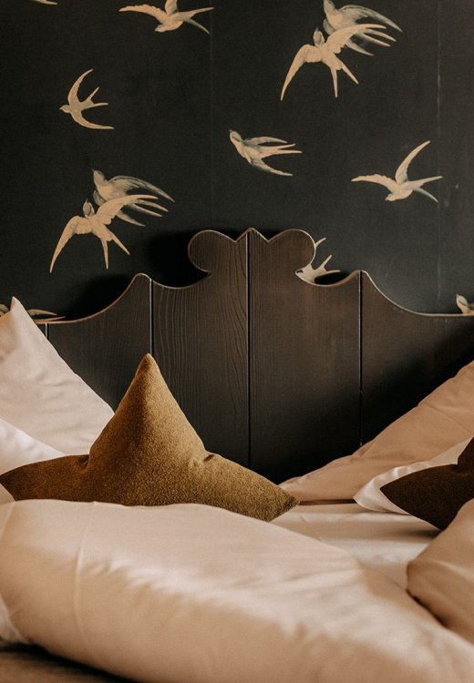 Close-up of a cozy bed with white bedding and an olive green upholstered headboard, with a swallow-patterned wallpaper.