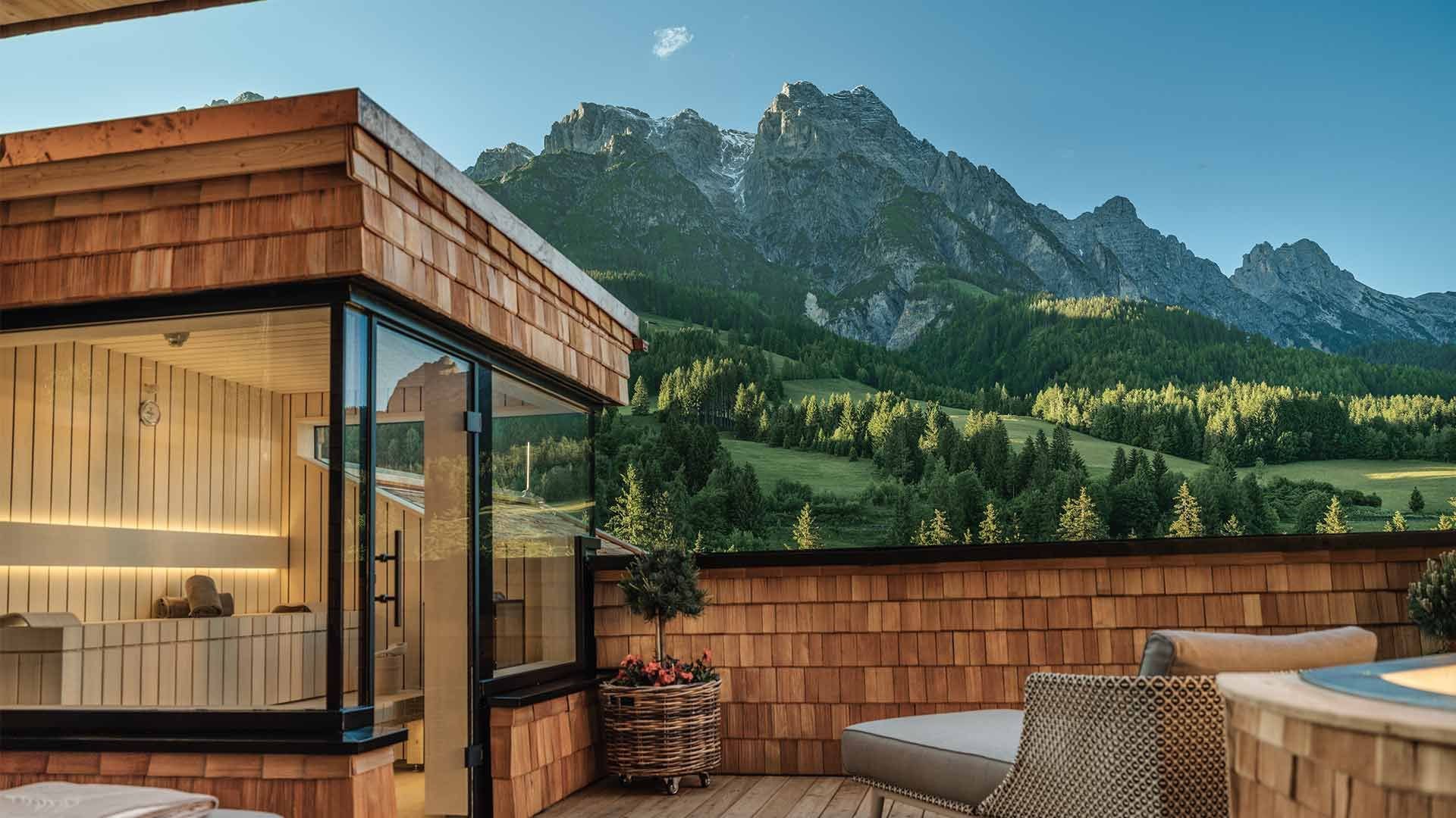 Exclusive rooftop terrace featuring a private wooden sauna, hot tub, and inviting seating areas, with impressive mountain views and vistas of lush alpine meadows. The panorama invites guests to relax and enjoy the beauty of the alpine nature.